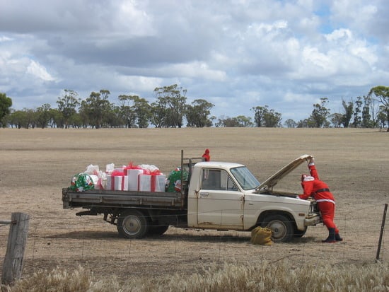 Papai Noel parado a frente de seu caminhão antigo com o capô aberto e com presentes na carroceria.